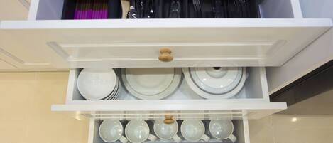 A kitchen cabinet with white shelves holding plates, bowls, and utensils.