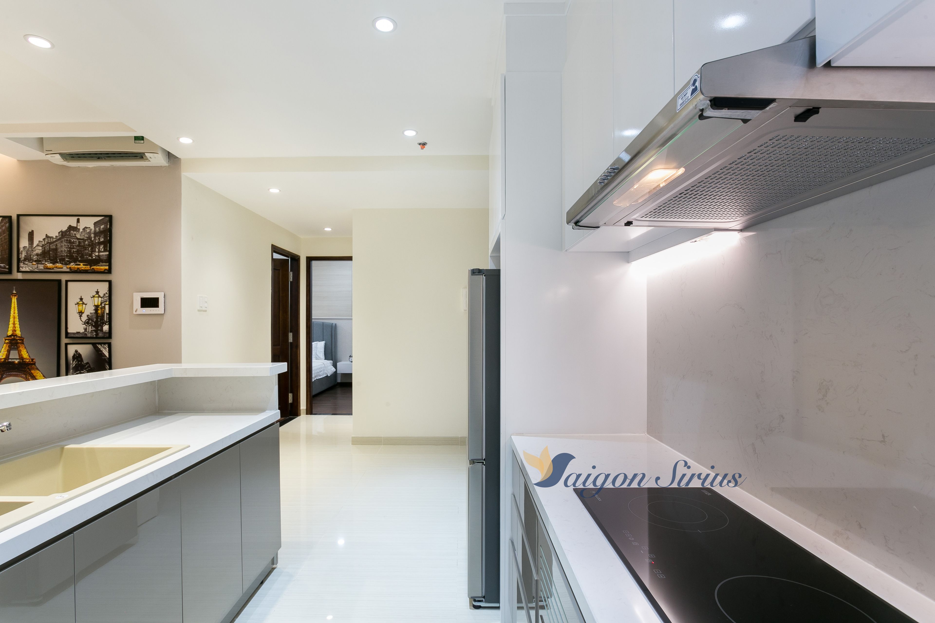 Une cuisine moderne équipée d’une cuisinière intégrée, d’un réfrigérateur et d’un évier. Des tableaux encadrés ornent le mur, et on peut apercevoir la chambre adjacente.