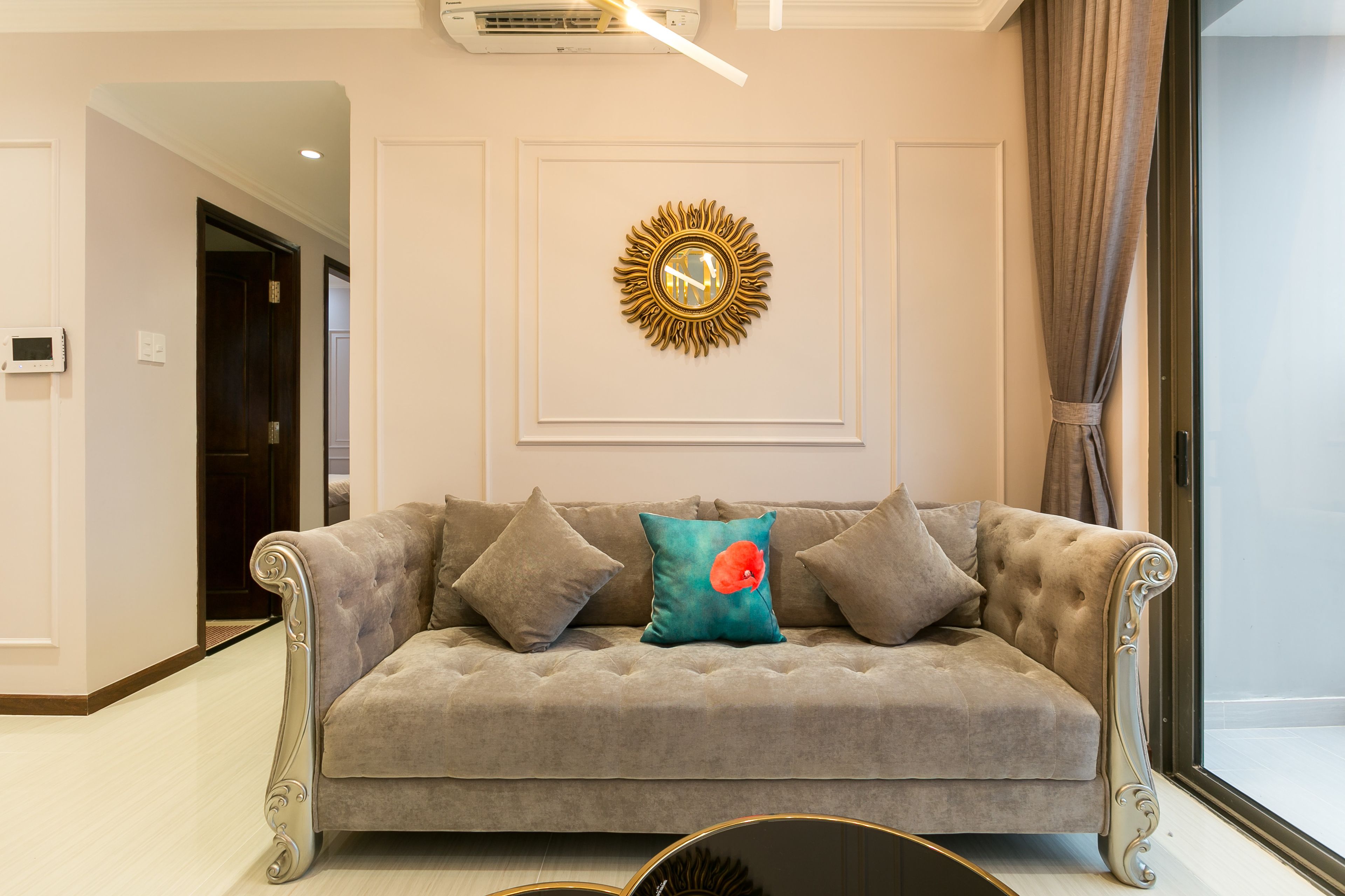 A plush grey sofa with decorative pillows, a wall-mounted sunburst mirror, and a large window with curtains.