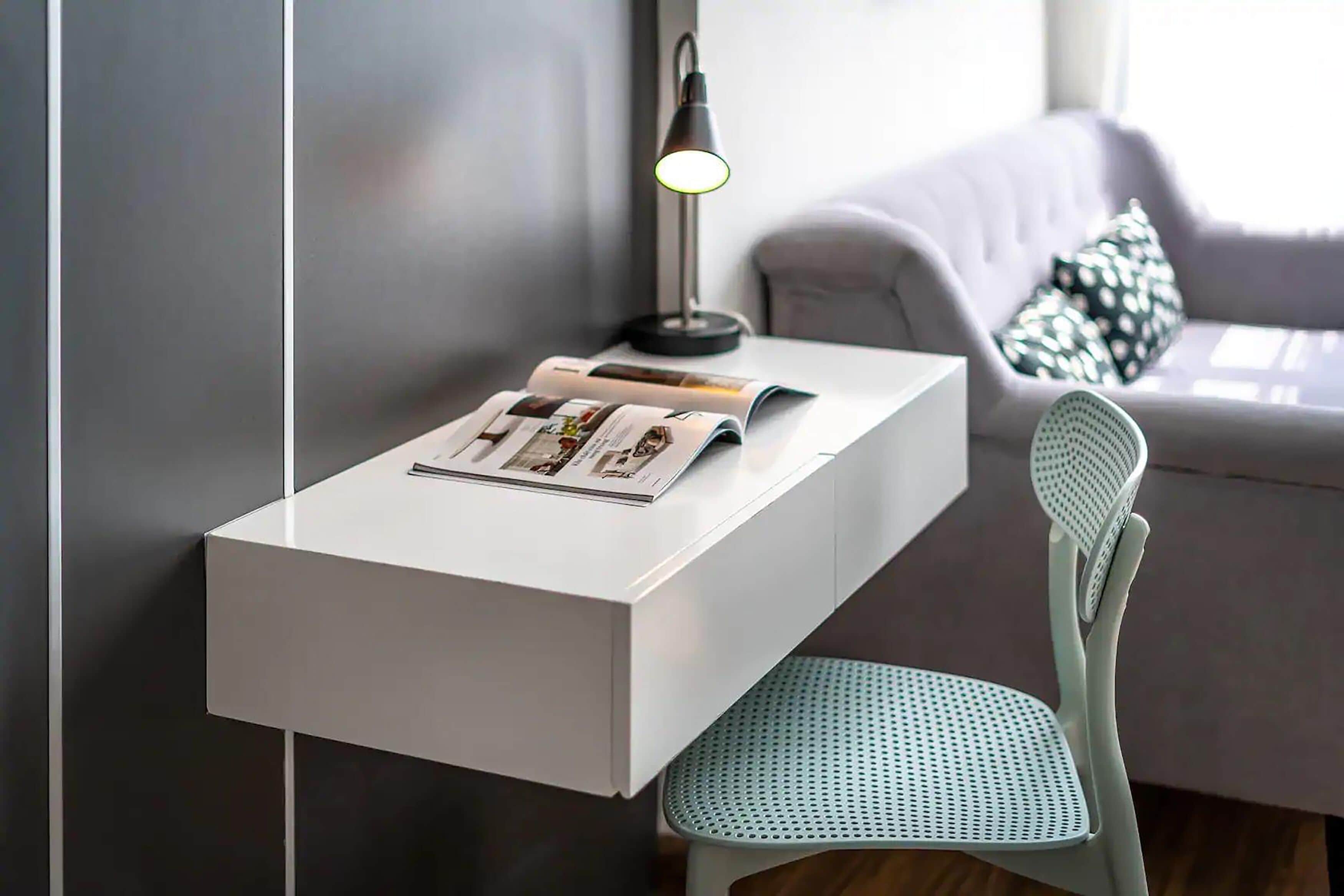 Et moderne værelse med et hvidt skrivebord, et magasin, en lampe og en grå sofa med mønstrede puder.