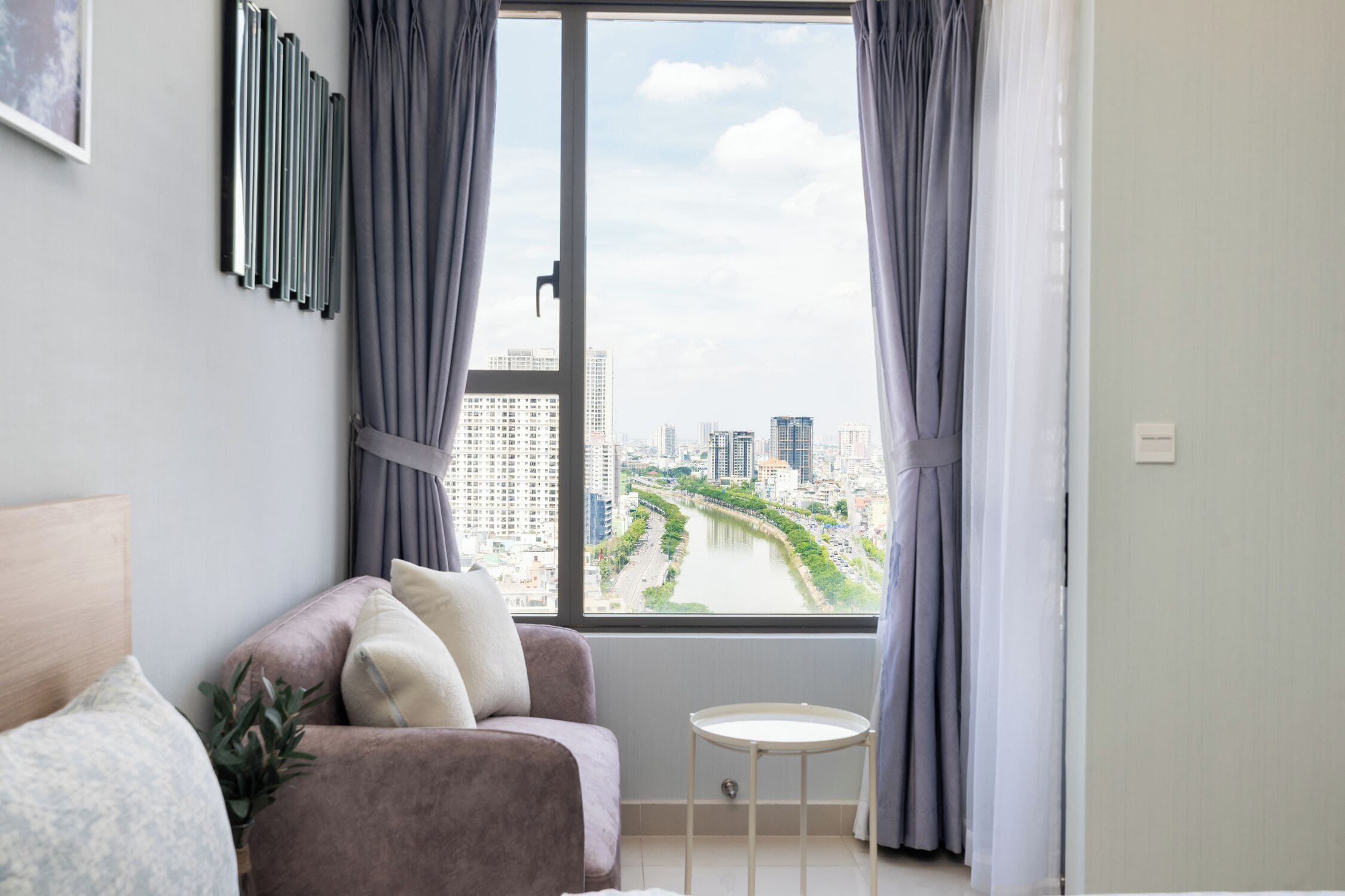 Una habitación moderna con vistas a un río y al paisaje urbano a través de amplios ventanales, una pequeña mesa redonda y una zona de estar con cojines.
