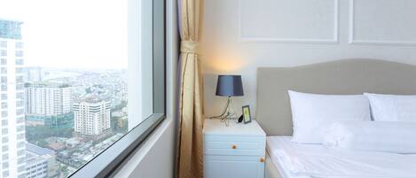 A hotel room with a bed, nightstand, lamp, and a view of a cityscape through a window.