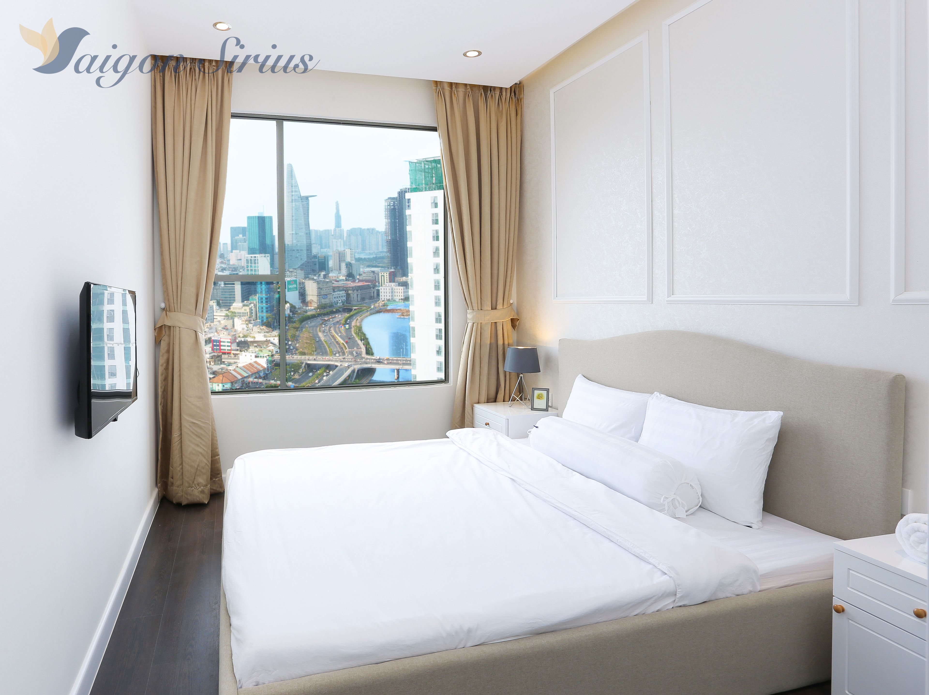 Una habitación de hotel con una cama, un televisor montado en la pared, una mesita de noche, una lámpara y una ventana con cortinas.