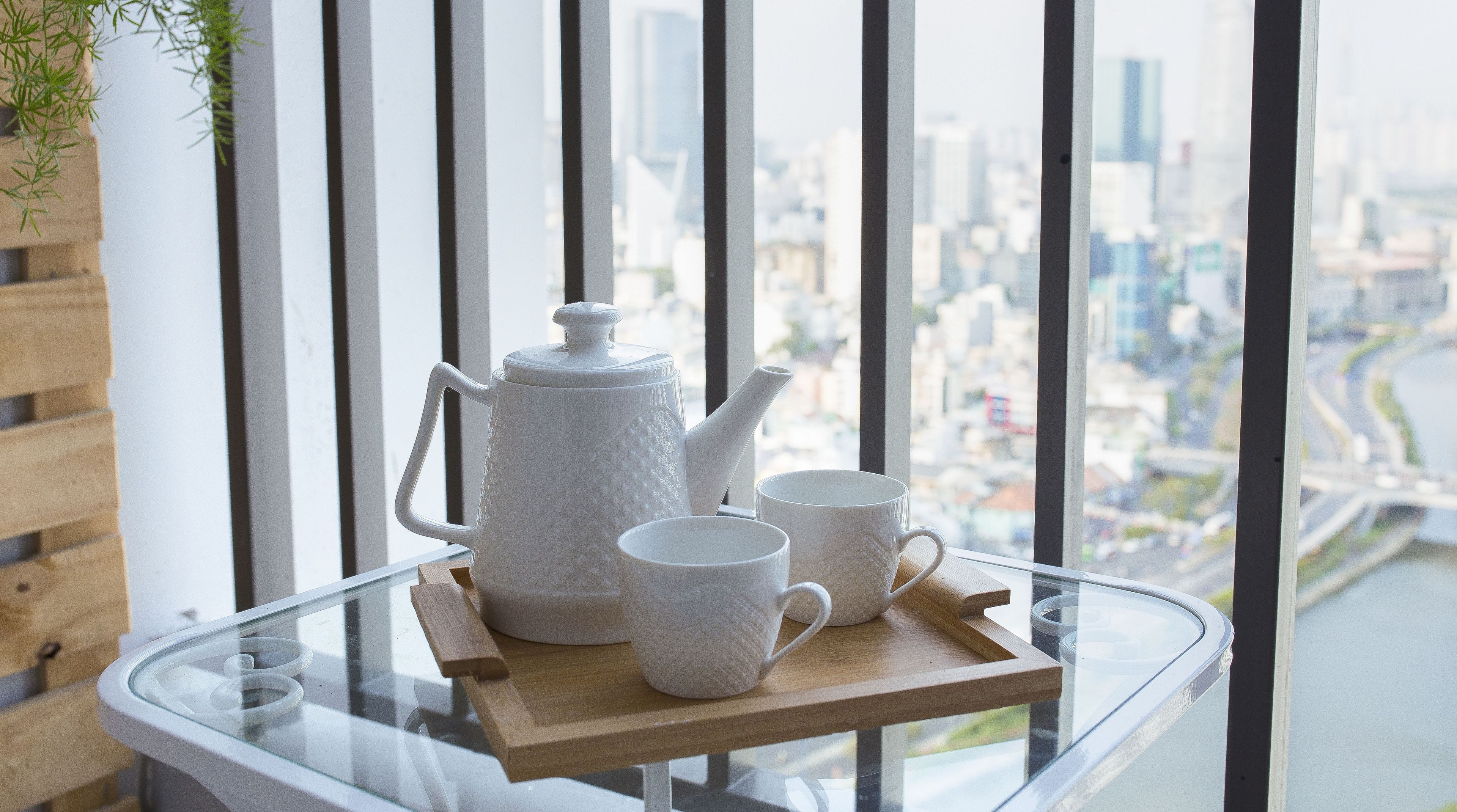 A glass table with a white teapot and two white cups on a wooden tray, overlooking a cityscape through a window with black vertical blinds.