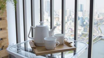A glass table with a white teapot and two white cups on a wooden tray, overlooking a cityscape through a window with black vertical blinds.