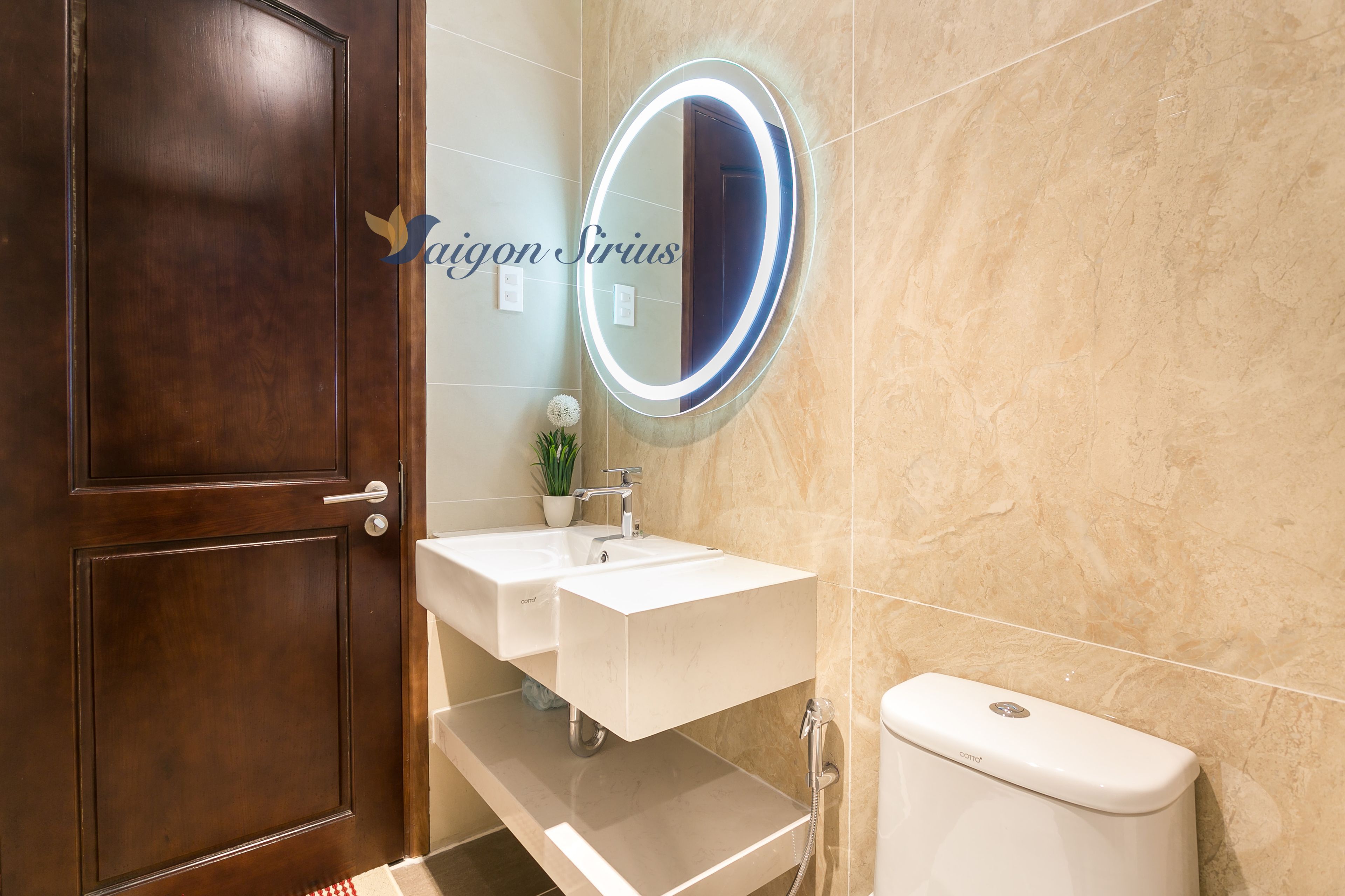 A bathroom with a wooden door, two sinks, a mirror with lighting, a toilet, and a potted plant.