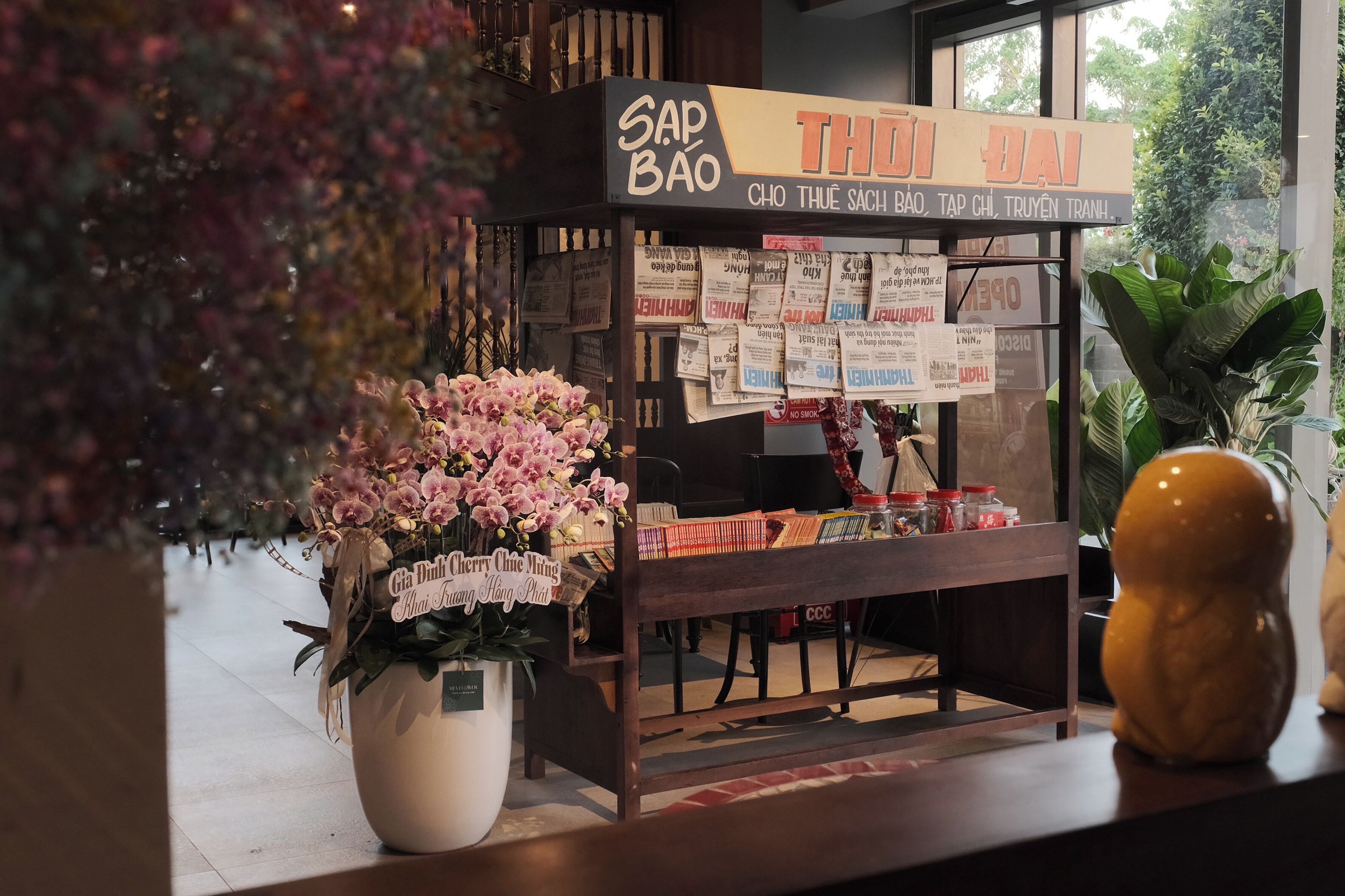 A bookstore with a sign reading "SAP BAO" and "THU DAO". There is a potted plant with pink flowers in the foreground.