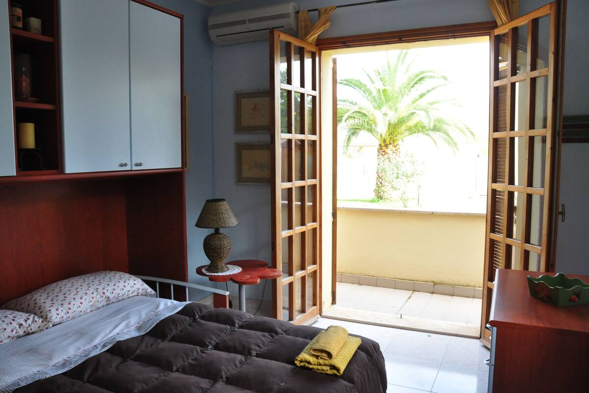 Une chambre avec un lit, une lampe, une étagère avec quelques objets et une vue sur un palmier, visible à travers une porte ouverte.