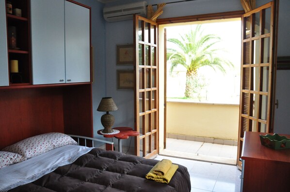 Un dormitorio con cama, lámpara, estante con objetos y vista a una palmera a través de una puerta abierta.