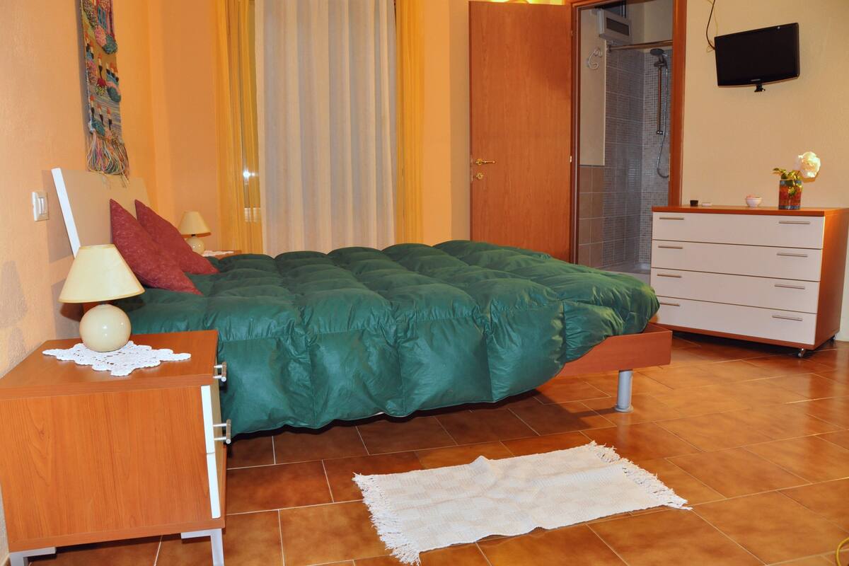 Une chambre avec un lit vert, une table de chevet en bois, une commode blanche et un téléviseur fixé au mur.