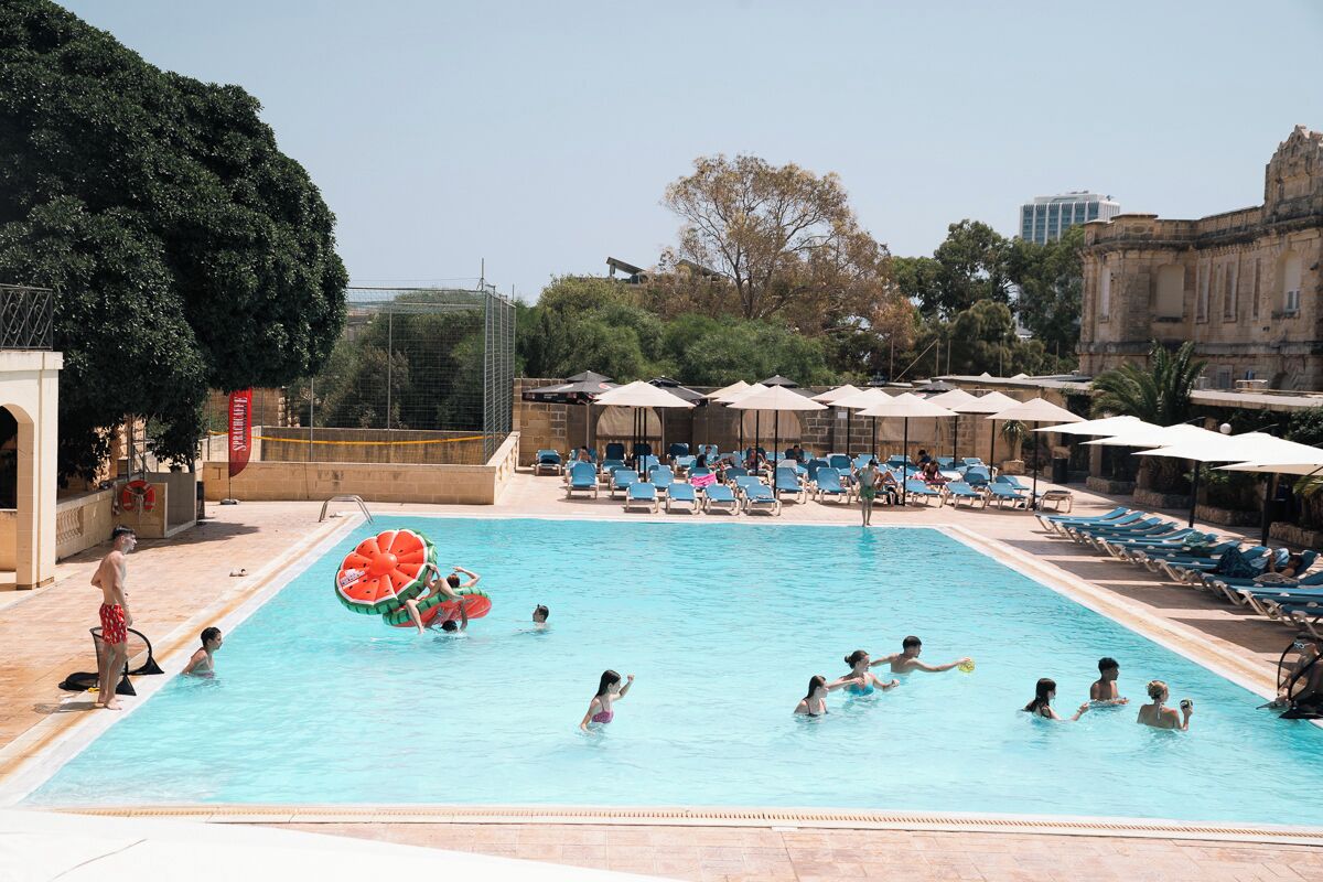 A swimming pool with lounge chairs and umbrellas, surrounded by trees and a building.