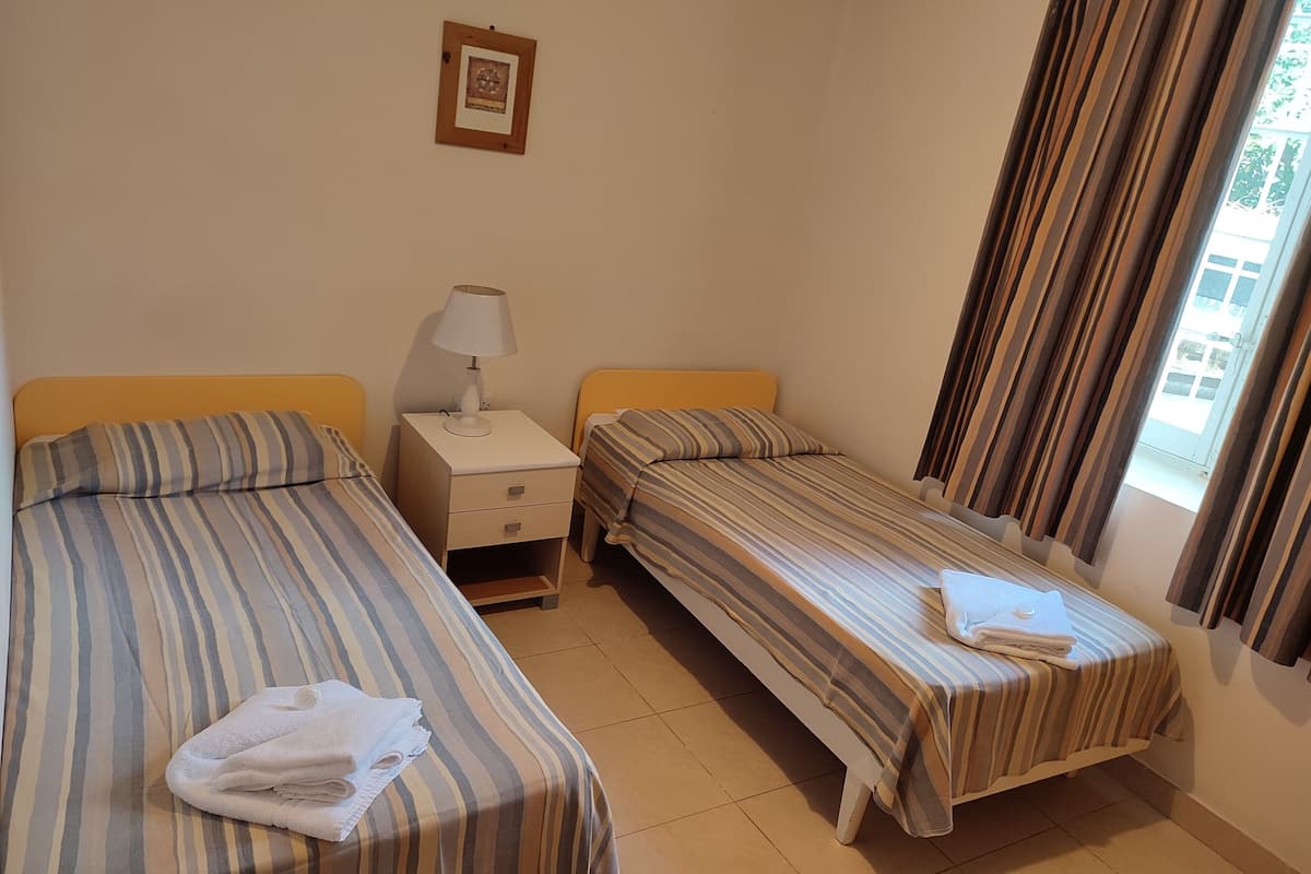 Habitación de hotel con dos camas individuales, una mesita de noche con lámpara, un cuadro en la pared y una ventana con cortinas.
