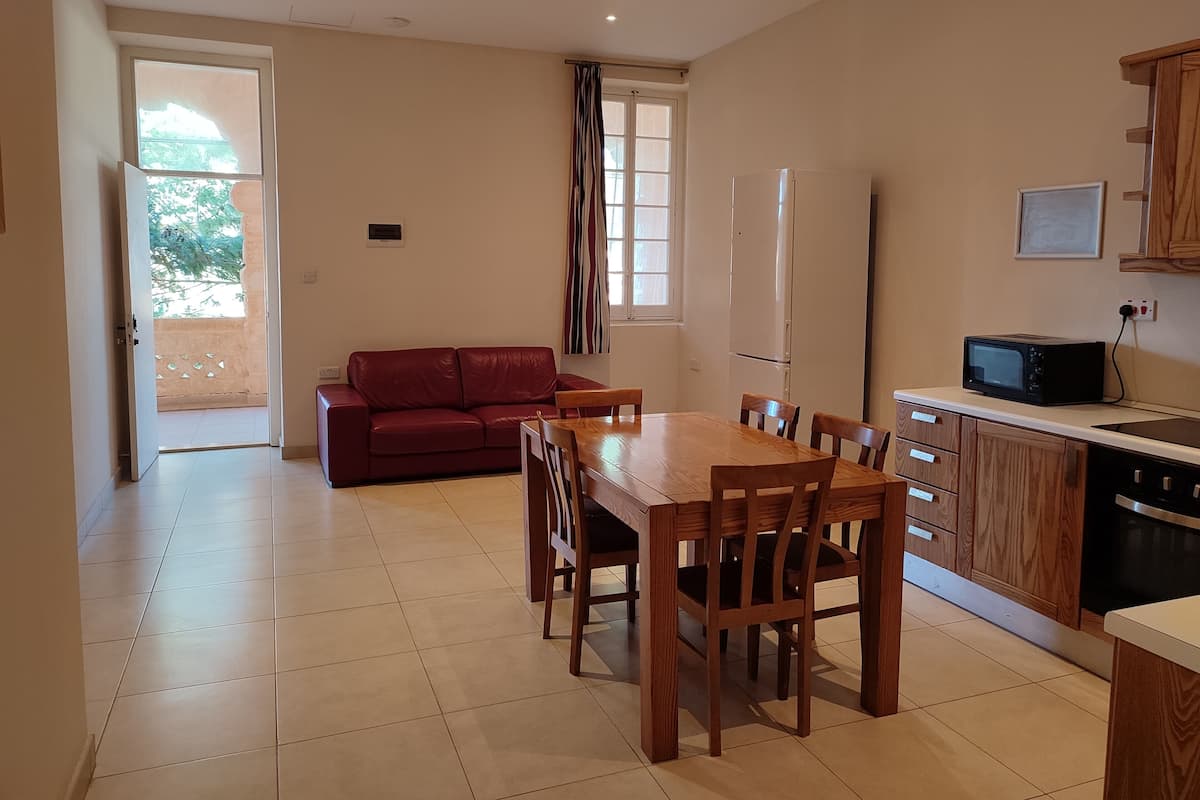 Amplia sala de estar con un sofá rojo, mesa de comedor de madera y sillas. Se observa una zona de cocina con microondas y armarios.