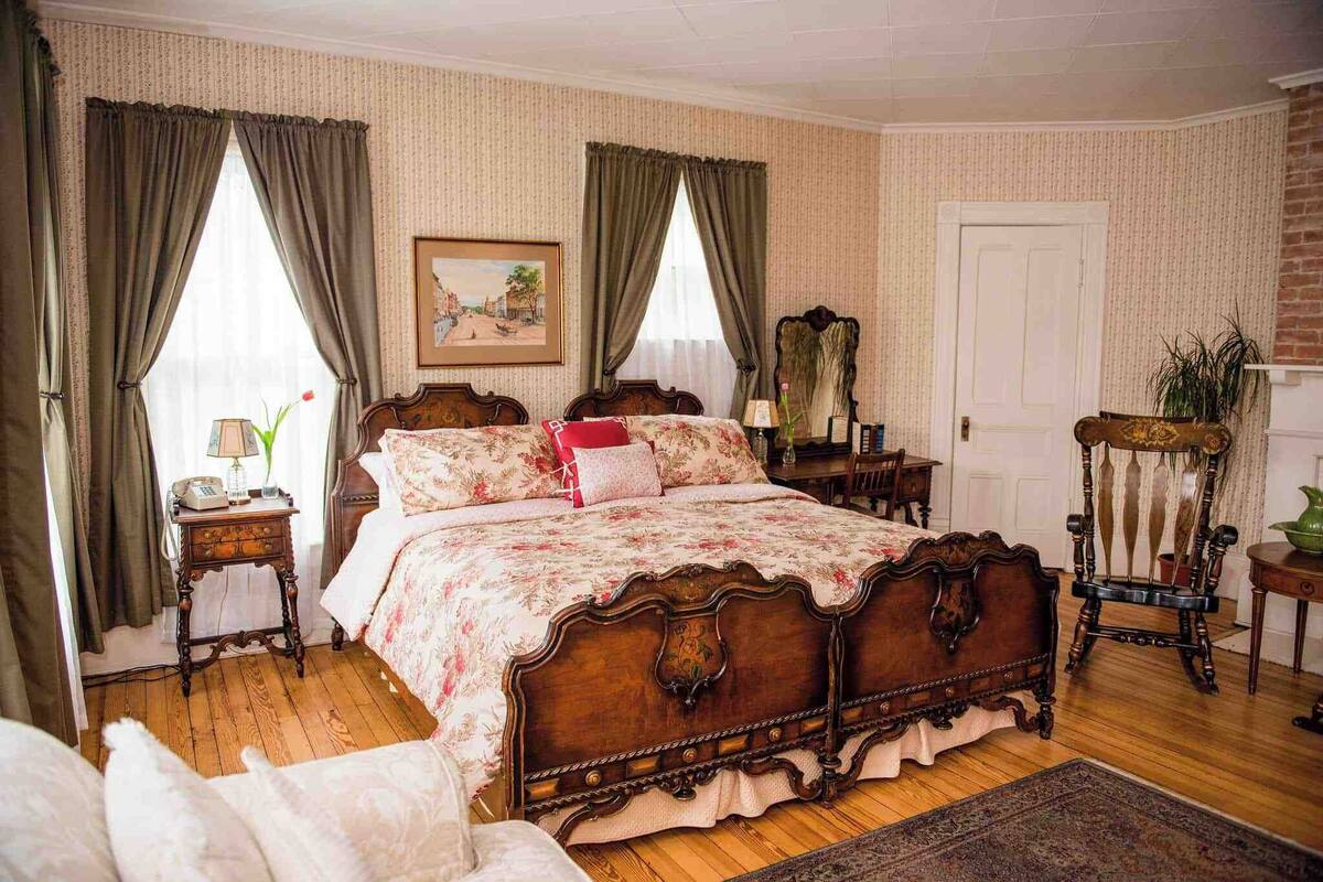 A traditional bedroom with a wooden four-poster bed, a nightstand with a lamp, a chair, and a framed picture on the wall.