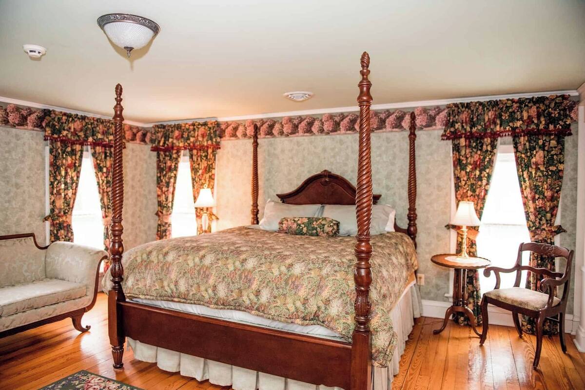 A four-poster bed with a floral bedspread, a wooden nightstand, a chair, and a small table with a lamp.