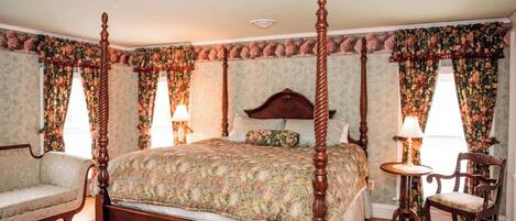 A four-poster bed with a floral bedspread, a wooden nightstand, a chair, and a small table with a lamp.