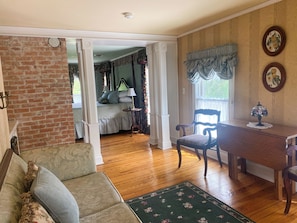 A room with a wooden floor, a green floral rug, a wooden desk, a chair, a bed with a green headboard, and a brick wall.