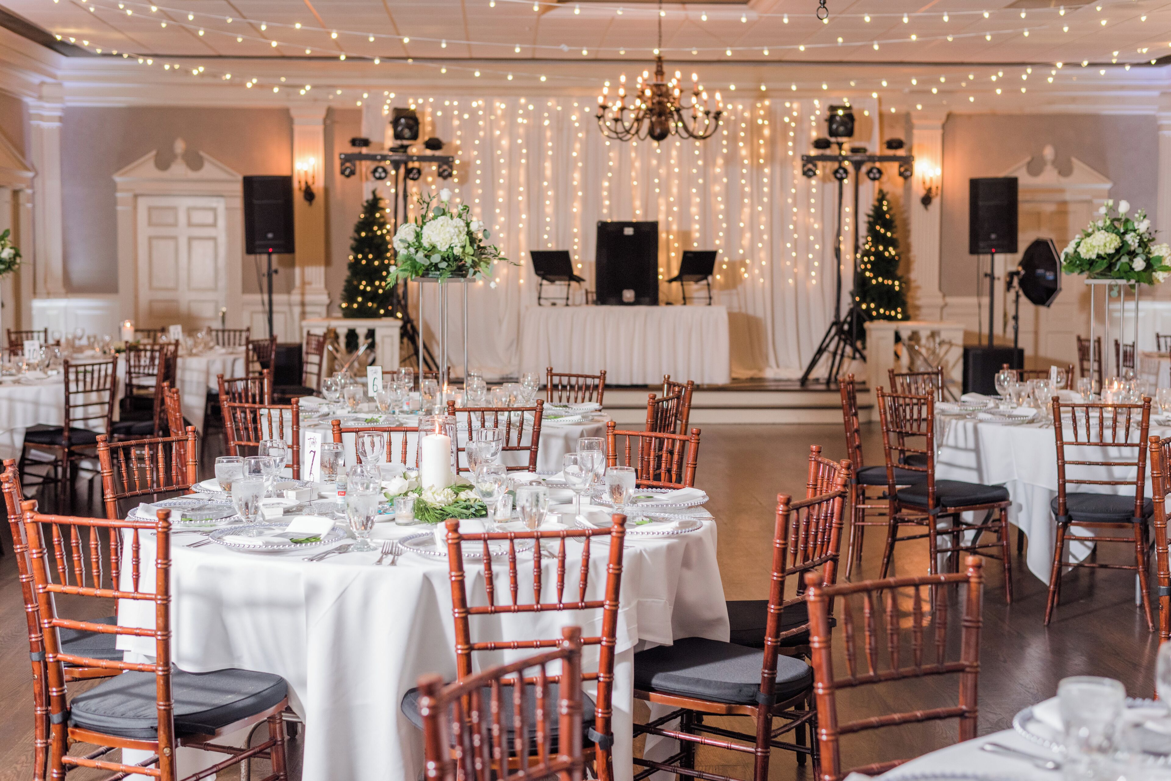 A banquet hall with round tables set for a formal event, featuring white tablecloths, wooden chairs, and a stage with musical instruments and a podium.