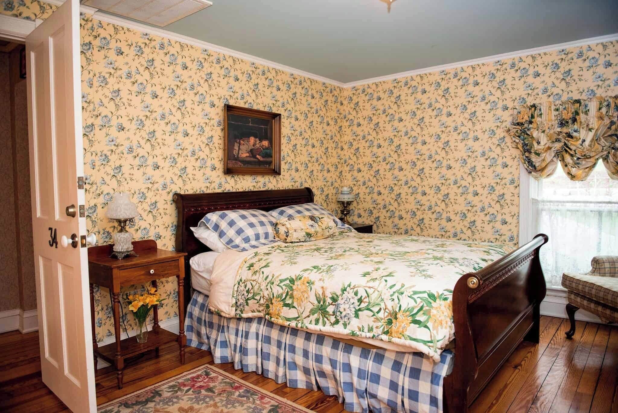 A bedroom with floral wallpaper, a wooden bed, a nightstand with a lamp, and a chair by the window.