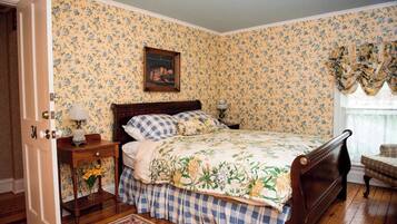 A bedroom with floral wallpaper, a wooden bed, a nightstand with a lamp, and a chair by the window.