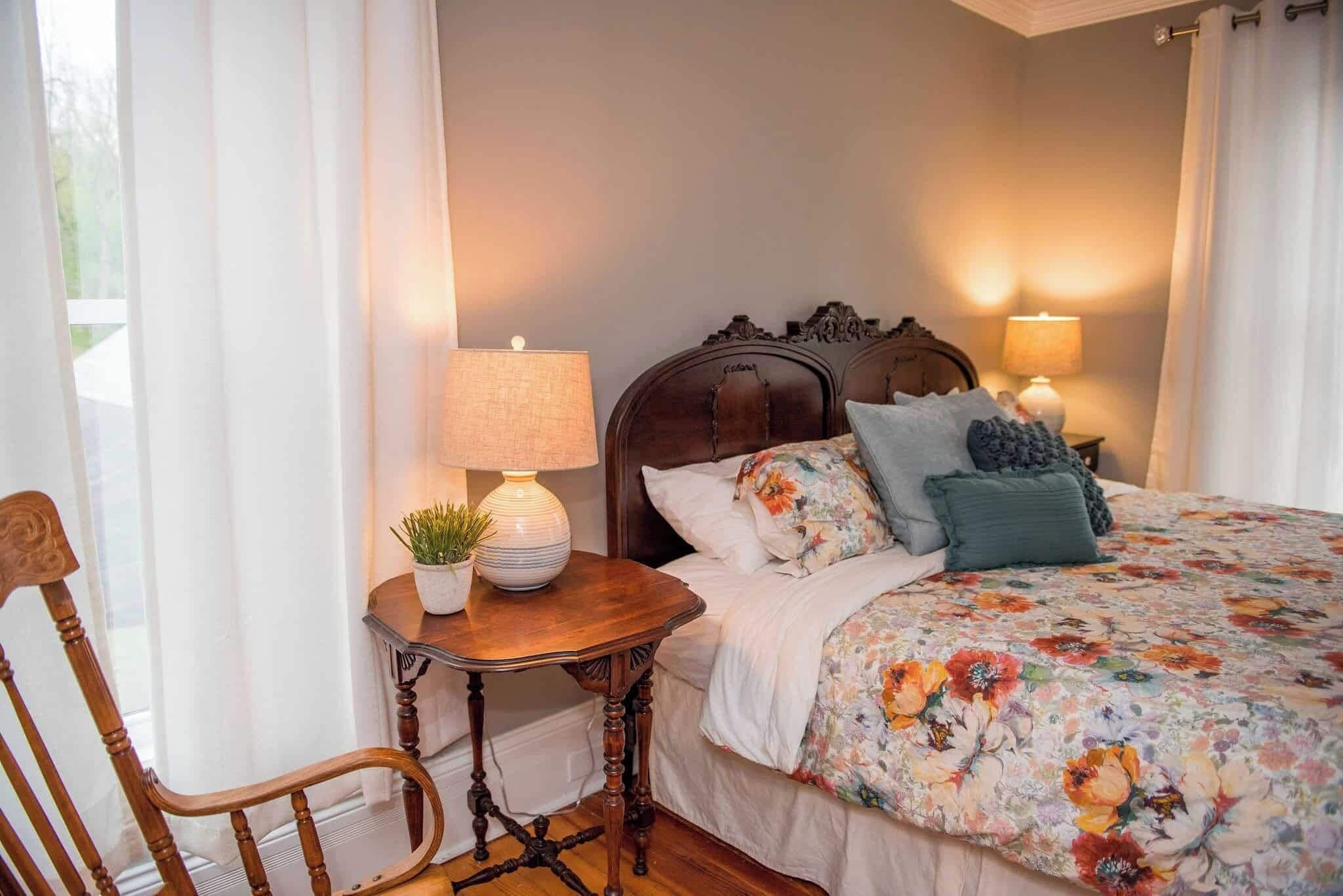 A bedroom with a wooden headboard, a floral bedspread, two bedside lamps, and a small table with a plant.