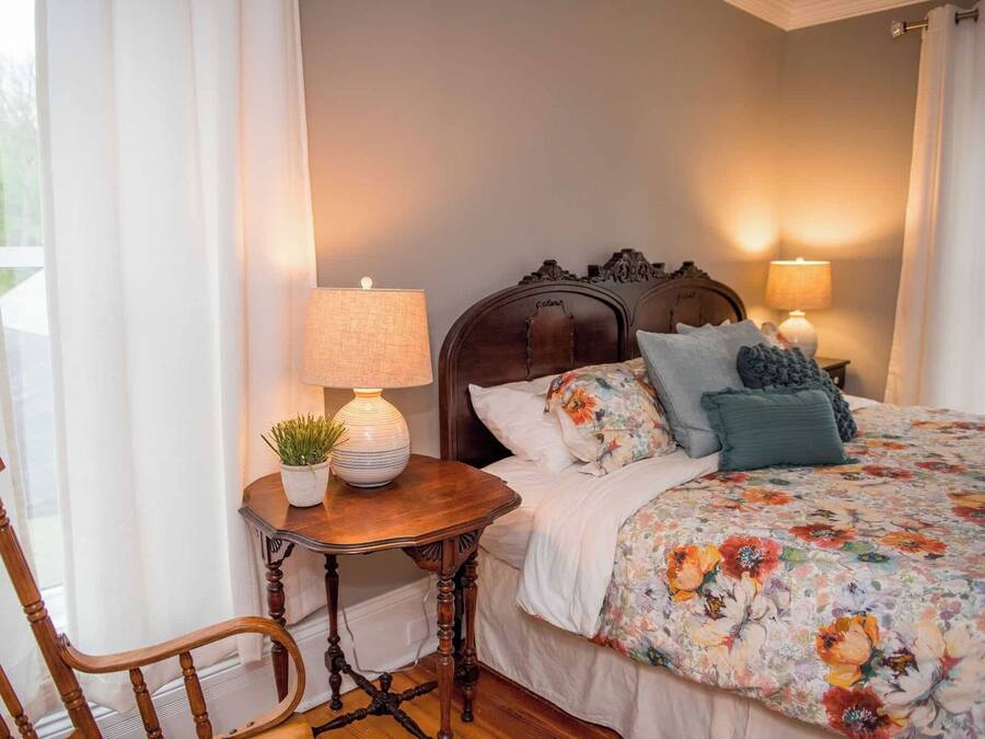 A bedroom with a wooden headboard, a floral bedspread, two bedside lamps, and a small table with a plant.