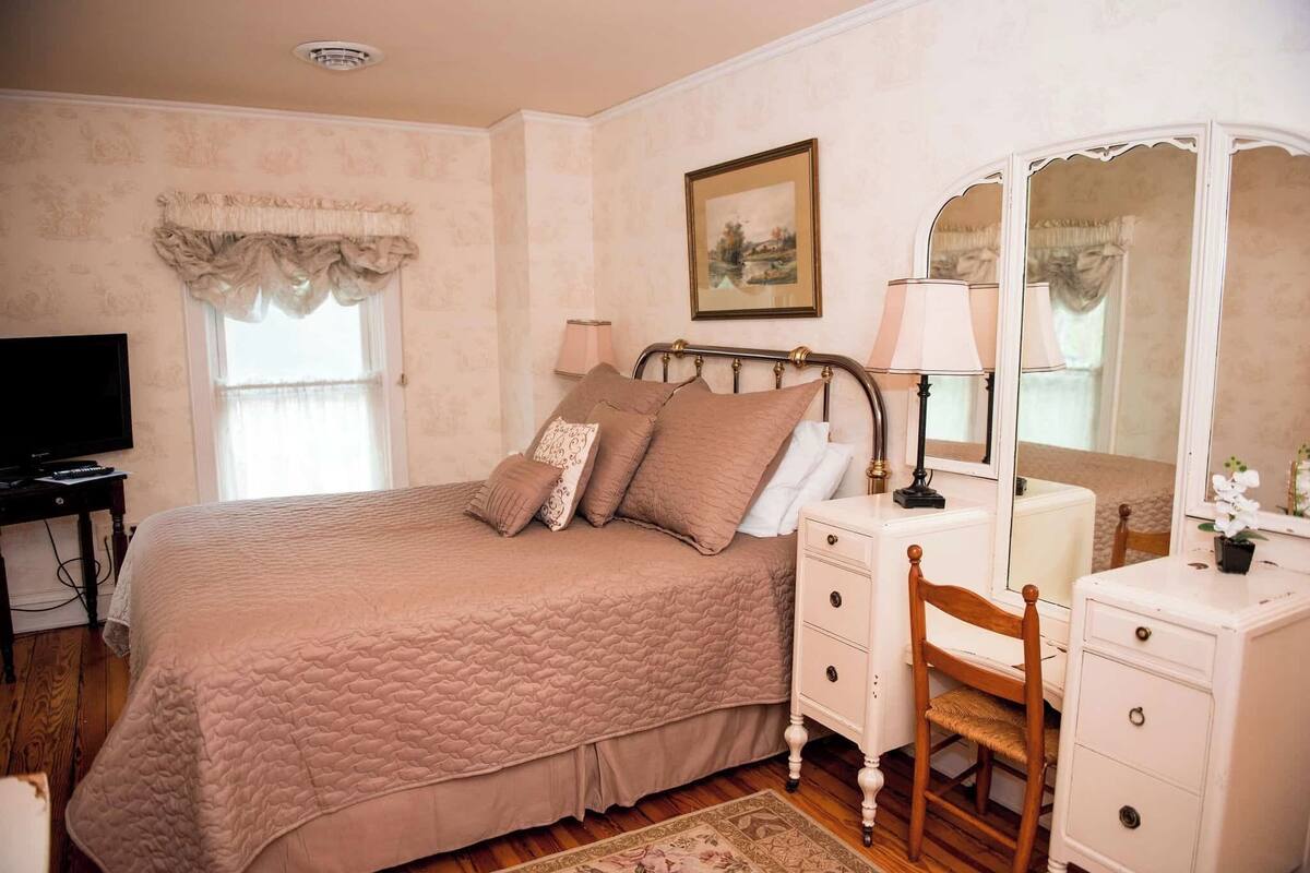 A bedroom with a bed, bedside table, lamp, dresser, mirror, and a painting on the wall.