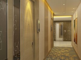 Um corredor com piso estampado geométrico, uma porta com placa de número e um aplique de parede.