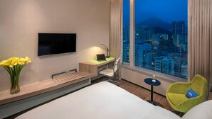 Un cuarto de hotel con cama, escritorio con laptop, televisión, silla, florero con flores y vista a la ciudad.