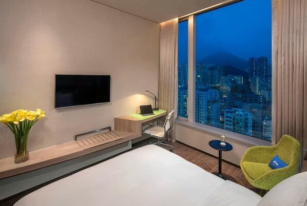 A hotel room with a bed, a desk with a laptop, a TV, a chair, a vase with flowers, and a view of a cityscape. - Holiday Inn Express Hong Kong Kowloon CBD2 by IHG (Kowloon)