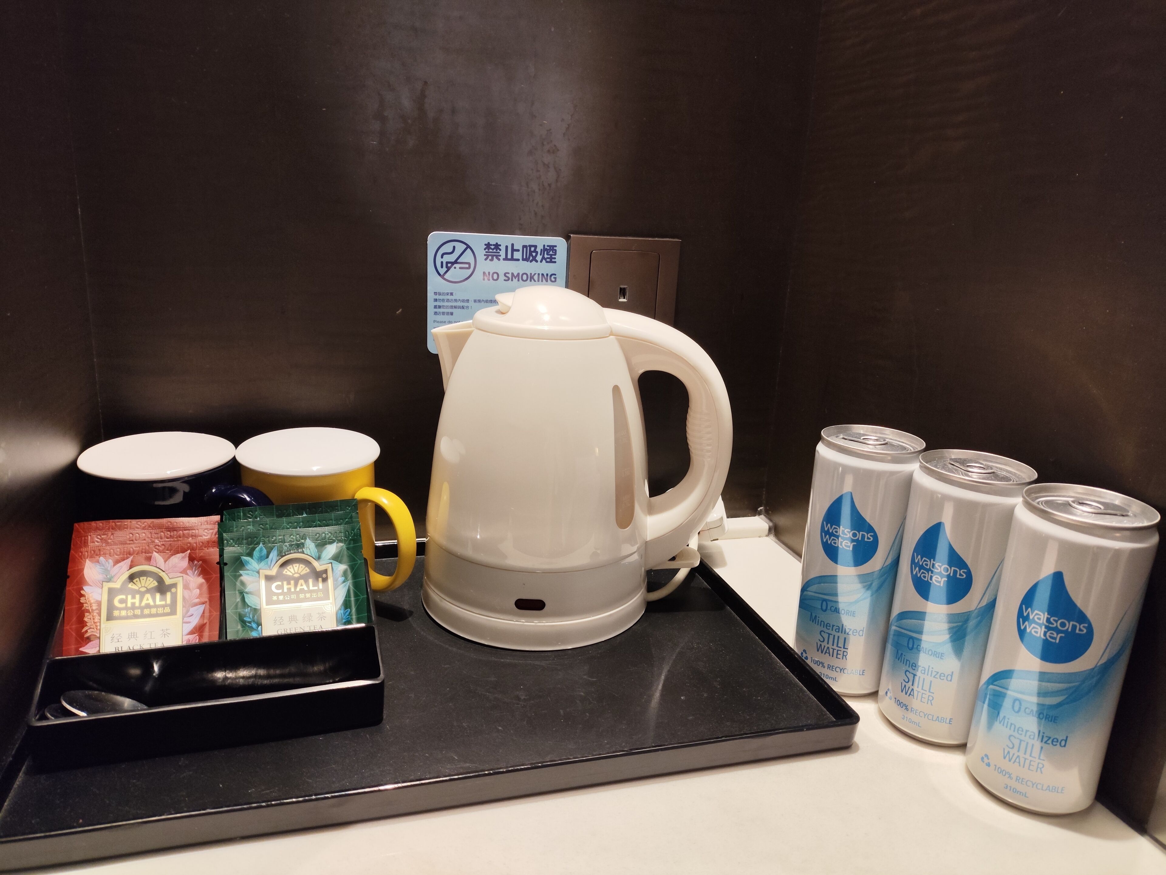A coffee station with a white coffee pot, two yellow mugs, and a tray with tea packets and cans of water.