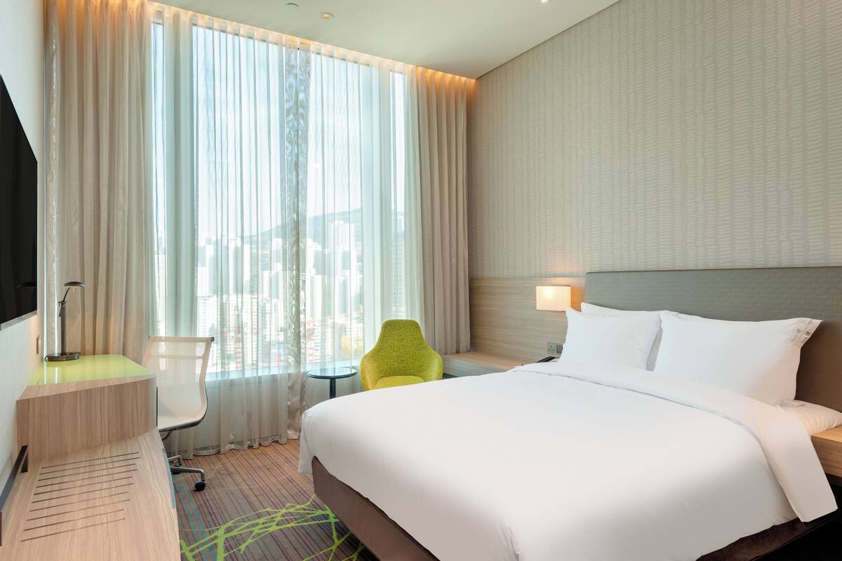 A hotel room with a large bed, a desk with a lamp, a chair, and a view of the city through sheer curtains.