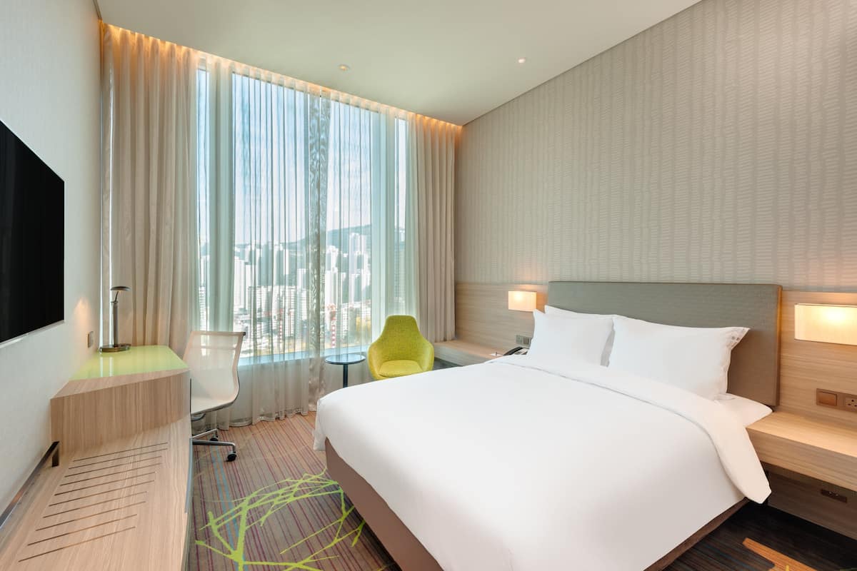 A modern hotel room with a large bed, a desk, a chair, and a view of the city through large windows.