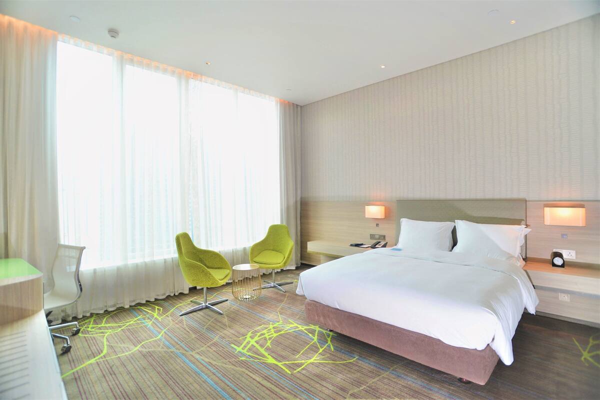 A hotel room with a large bed, two chairs, a desk, and a window with sheer curtains.