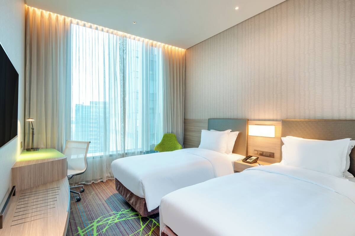 A hotel room with two beds, a desk, a chair, a lamp, and a view of the city through large windows.