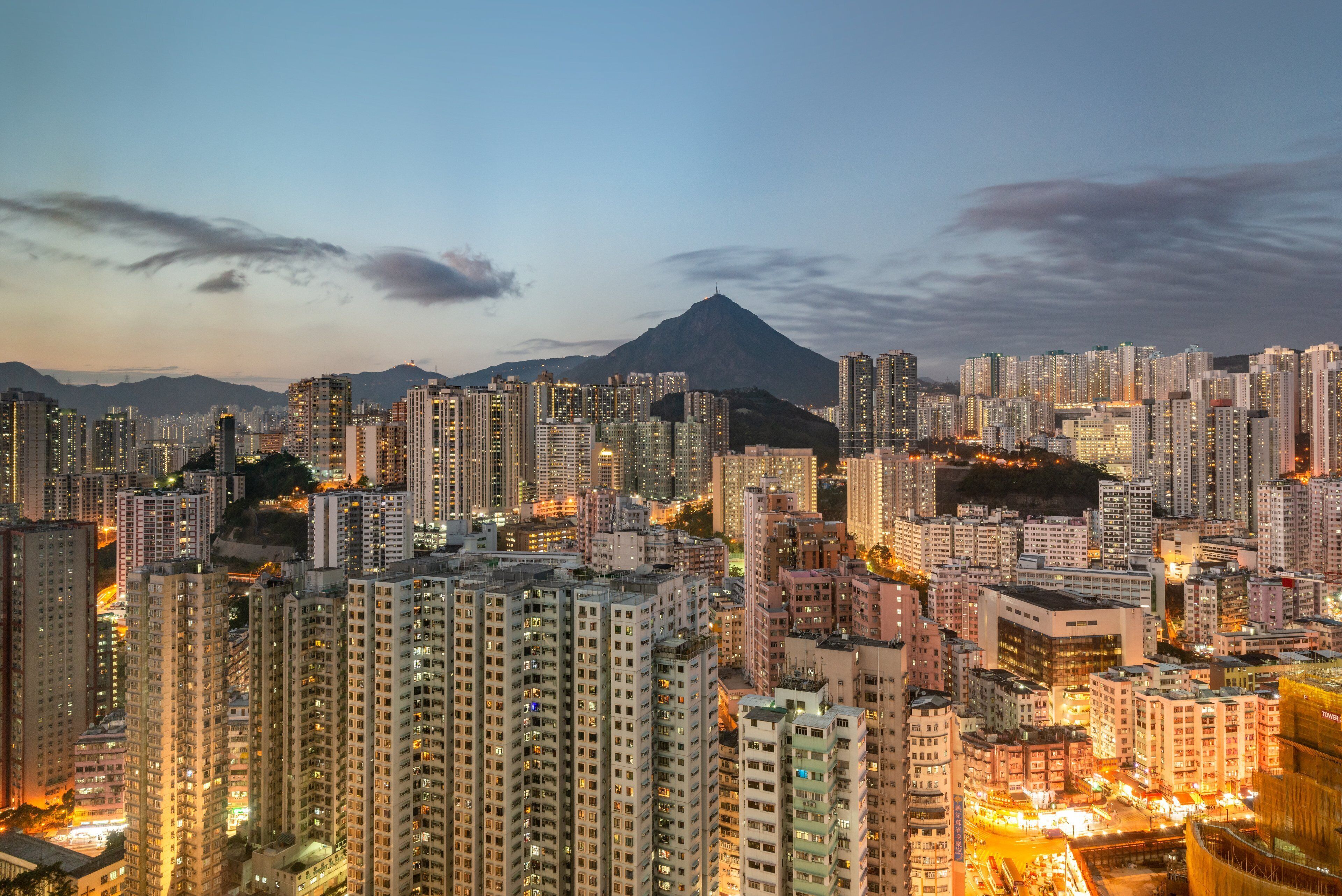 Céu urbano denso com arranha-céus, uma montanha imponente ao fundo e uma rua bem iluminada abaixo.