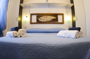 Una cama bien tendida con un edredón azul acolchado, dos toallas enrolladas y un cuadro en la pared.
