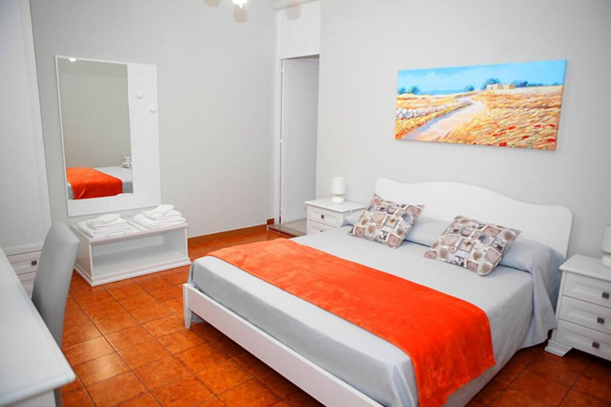 A bedroom with a bed, a bedside table, a nightstand, a mirror, a dresser, and a painting on the wall.