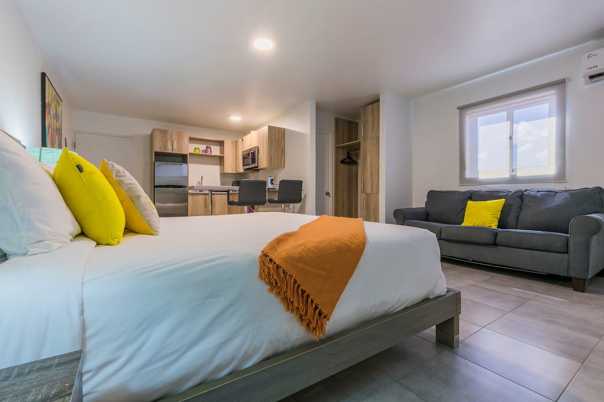 Um quarto com uma cama, um sofá cinzento e uma cozinha ao fundo.