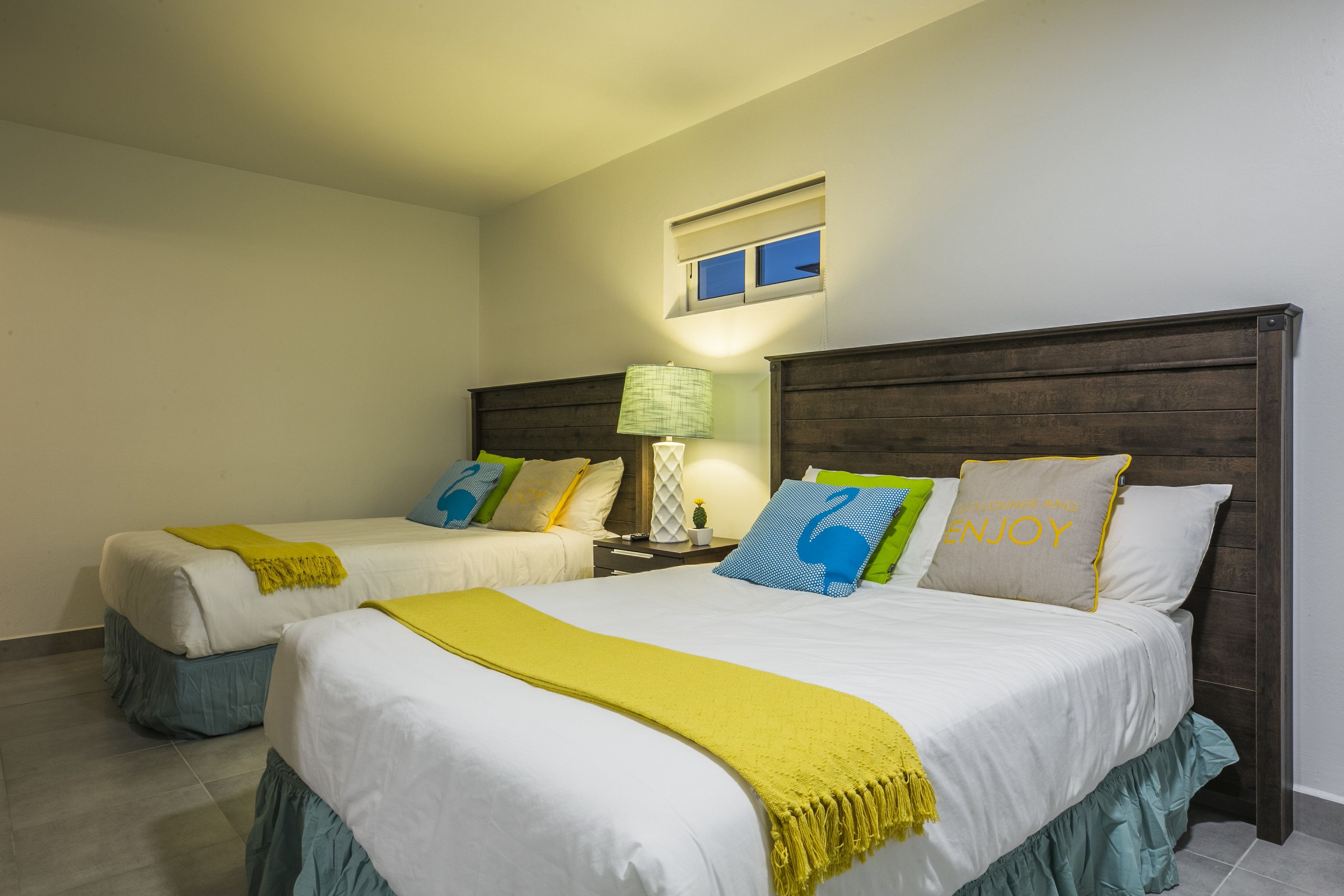 A hotel room with two beds, each with a yellow throw blanket, a nightstand with a lamp, and a window with blinds.