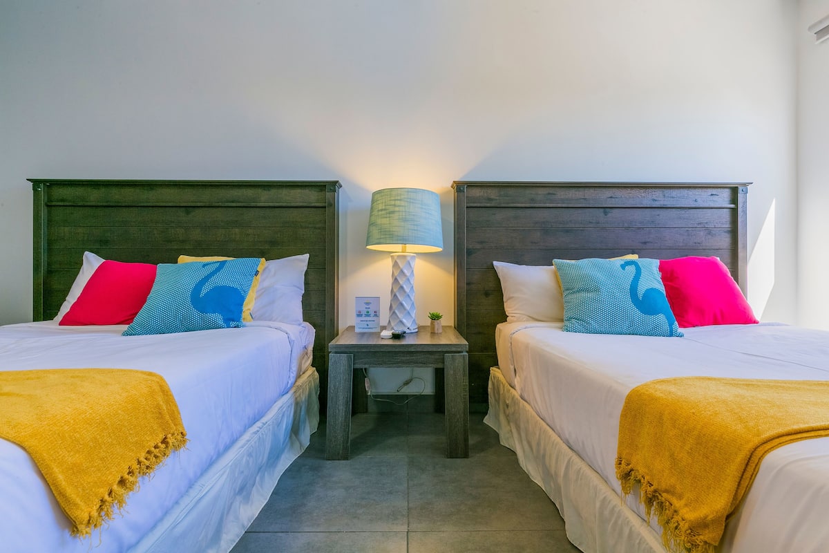 Quarto duplo com duas camas, cada uma com roupa de cama branca e um cobertor amarelo. Há uma mesa de cabeceira entre as camas, com uma luminária e uma pequena planta.