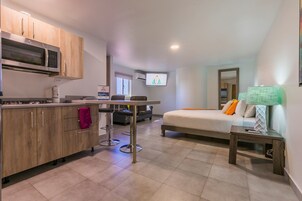Cocina compacta con microondas, armarios de madera y un taburete de bar. En la habitación contigua se ve una cama con almohadas y una mesita de noche con lámpara.