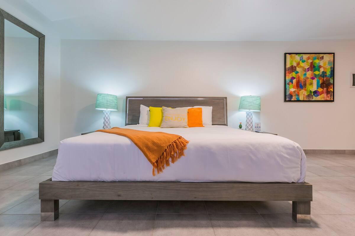 Quarto de hotel moderno com uma cama grande, duas luminárias de cabeceira, um quadro abstrato colorido e um espelho grande.