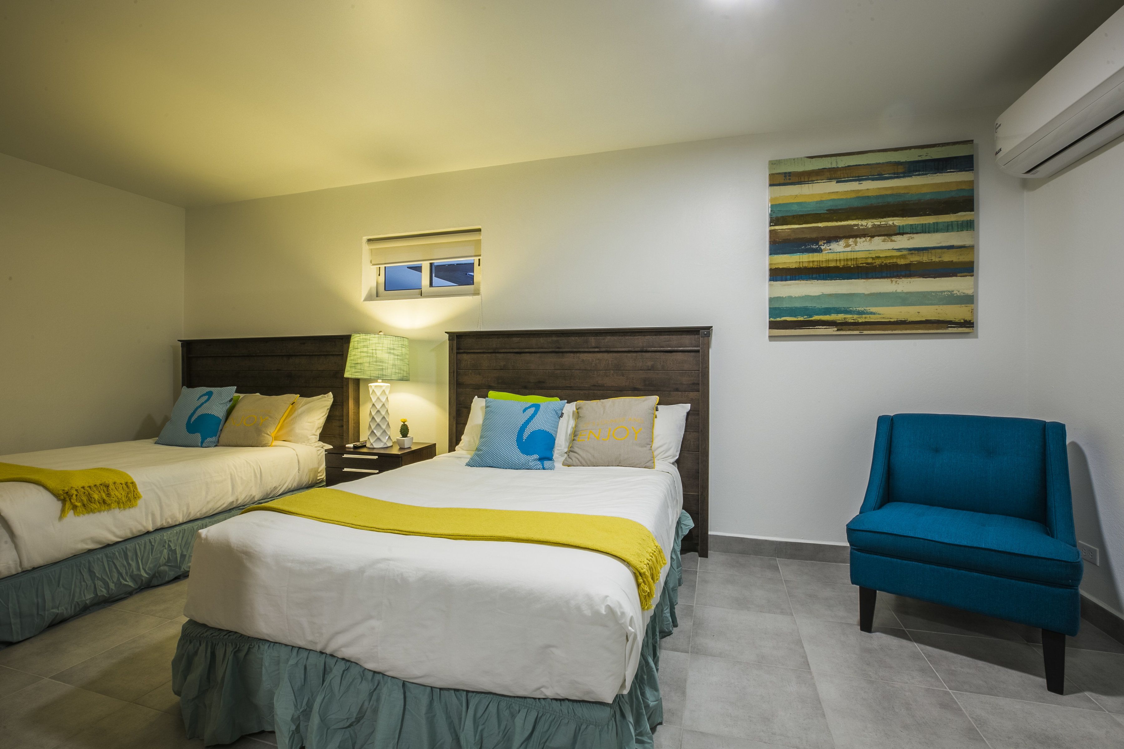 A hotel room with two beds, a nightstand, a lamp, a blue armchair, and a wall art piece.