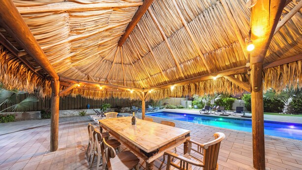 An outdoor dining area with a thatched roof, a pool, and a wooden table with chairs. - Turibana Plaza (Noord)