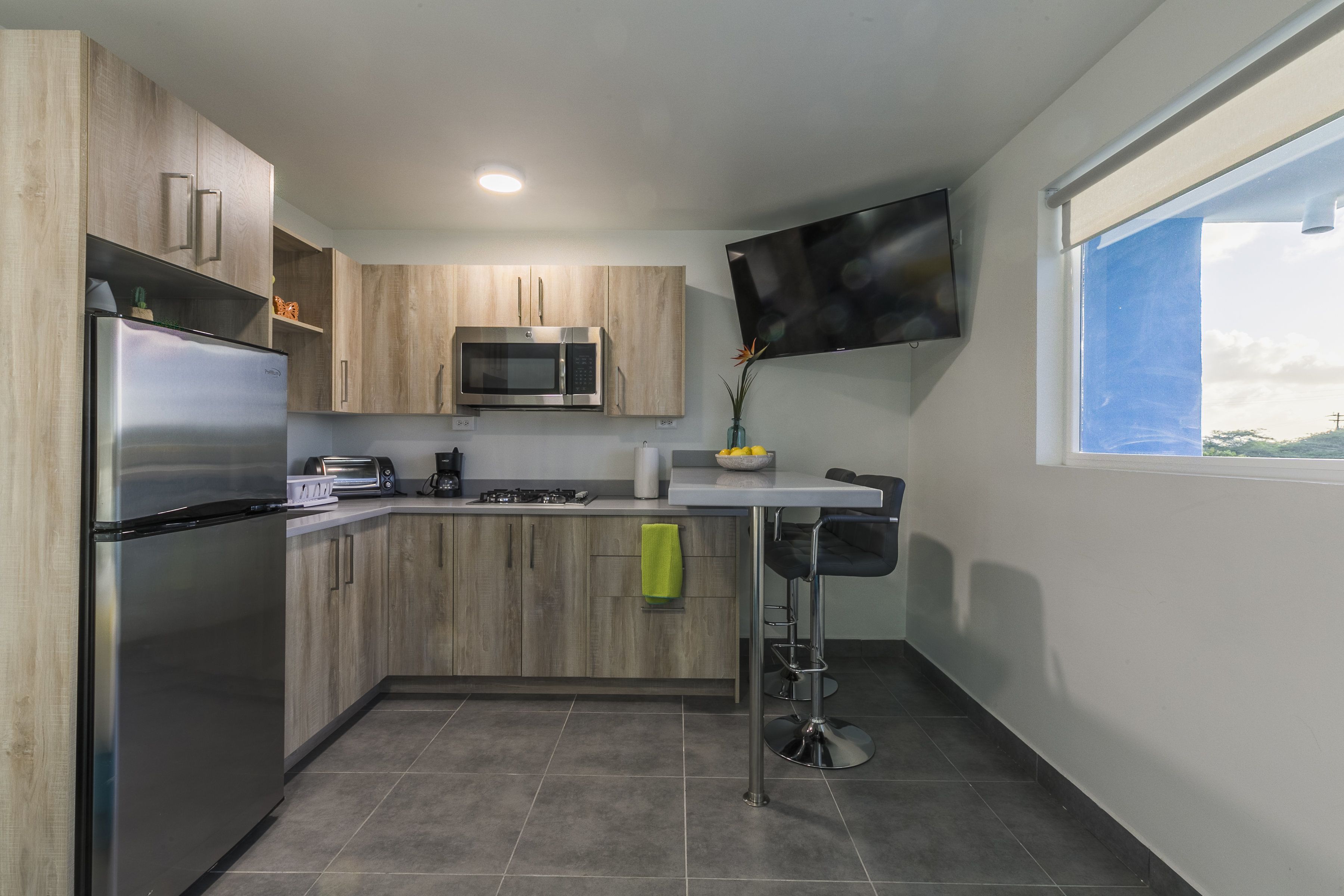 A modern kitchen with wooden cabinets, a stainless steel refrigerator, a microwave, and a flat-screen TV mounted on the wall.