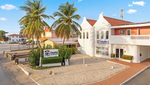 A building with a sign for Vacation Aruba.com, surrounded by palm trees and a gravel area. - Turibana Plaza (Noord)