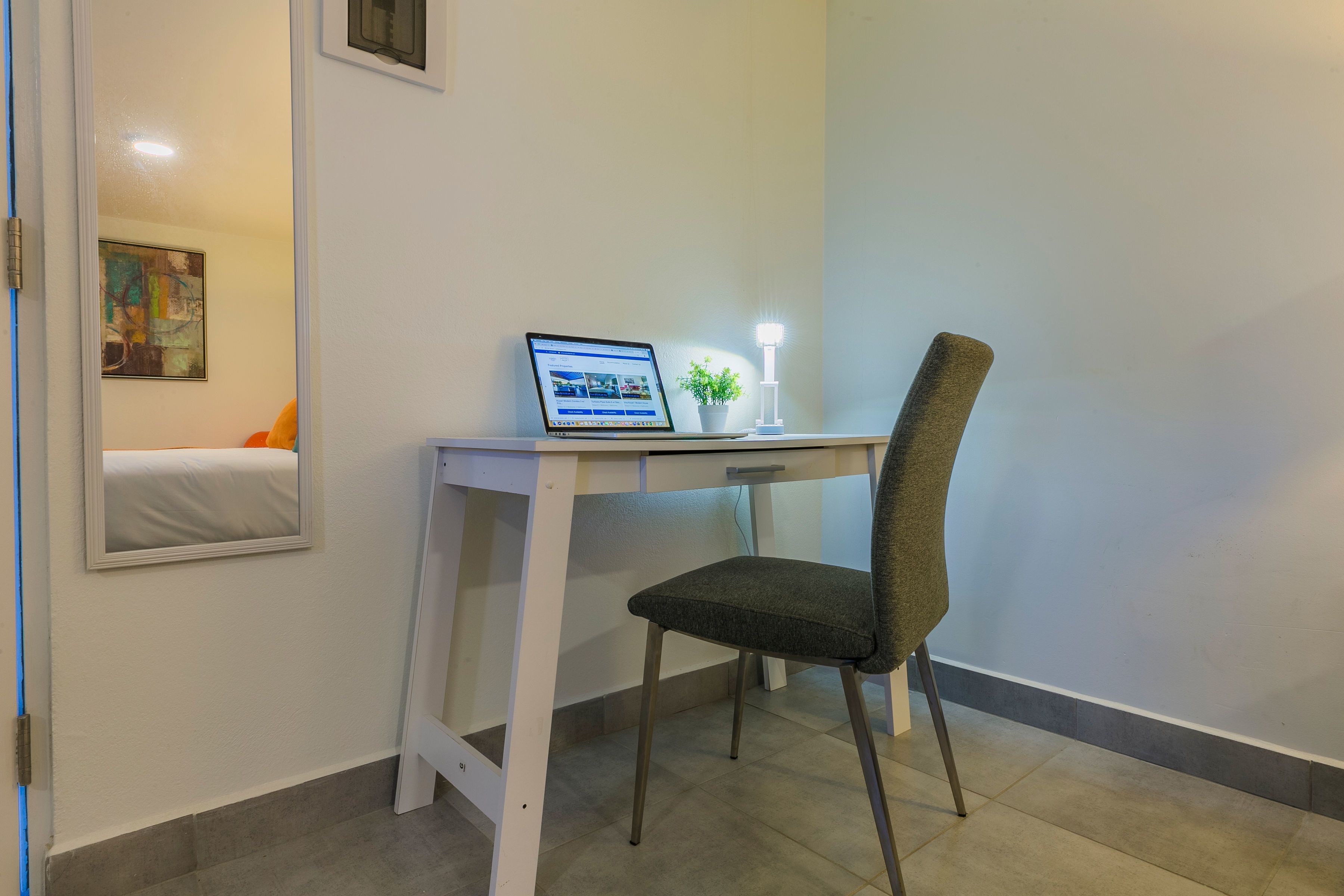 A hotel room with a desk, chair, laptop, bedside table, lamp, and a painting on the wall.