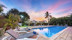 A swimming pool with lounge chairs, surrounded by palm trees and a wooden fence. - Turibana Plaza (Noord)