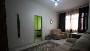 Ein Wohnzimmer mit einem beigen Sofa, einer grünen Tür, die in einen anderen Raum führt, einem Deckenventilator und einem Fenster mit Vorhängen.